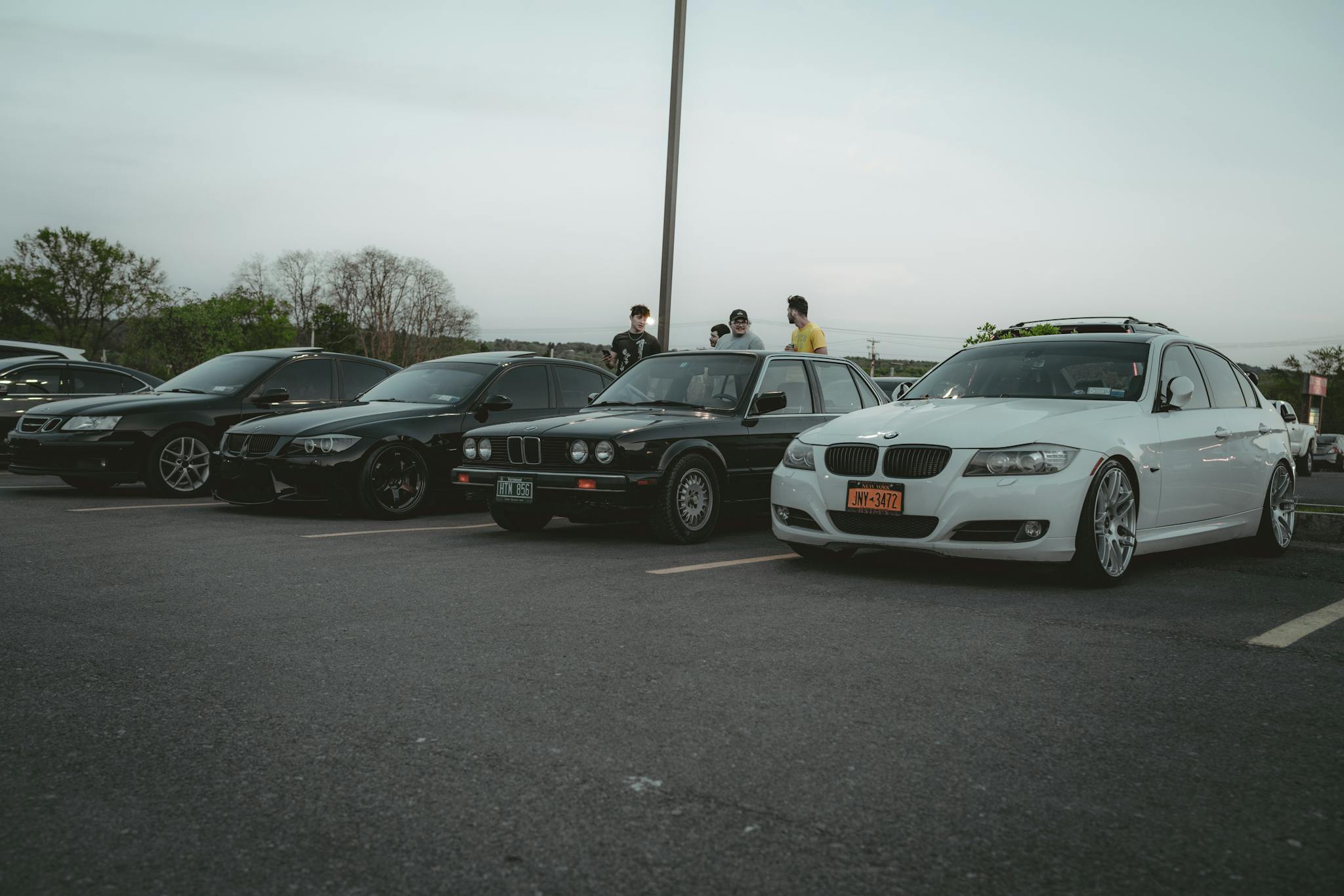 A lineup of BMW cars parked in an outdoor lot with people gathering during the evening.