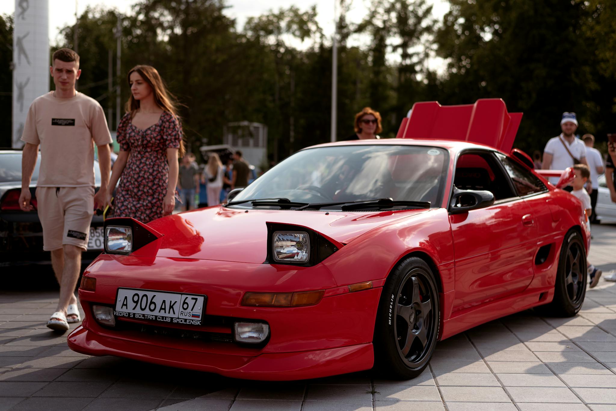 A young couple walking past a red Toyota MR2 on a sunny urban day, creating a vibrant lifestyle scene.