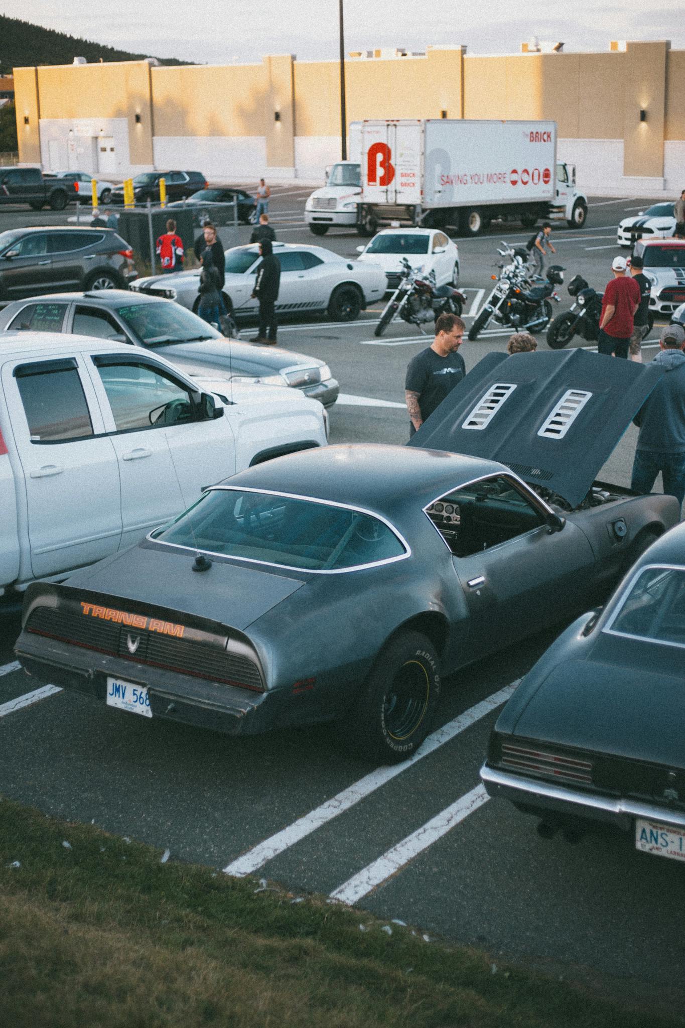 Outdoor car meet featuring classic vehicles, people, and motorbikes on a sunny day.